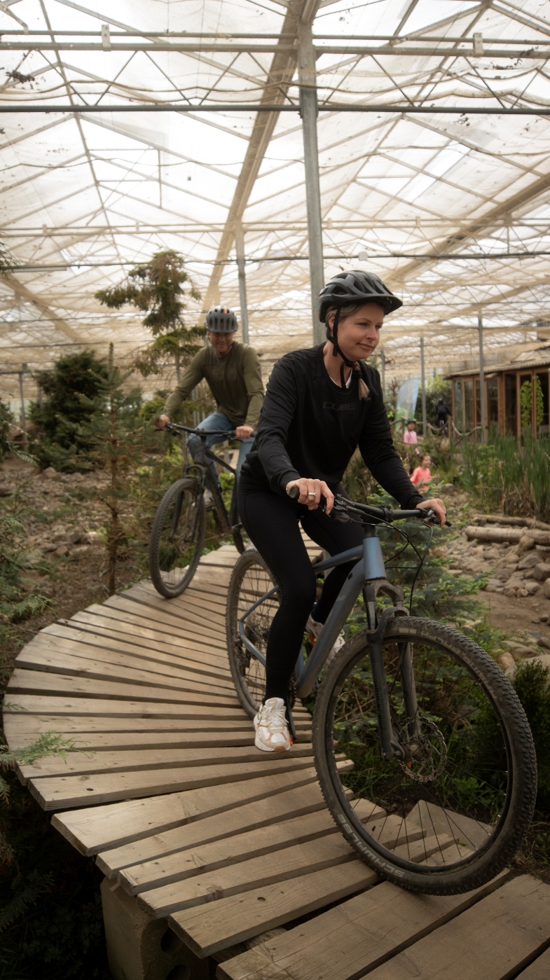 Twee volwassenen mountainbiken over de houten handbrug op het overdekte indoor parcours van Indoor Mountainbike Almere, tussen het groen onder het lichte kasdak