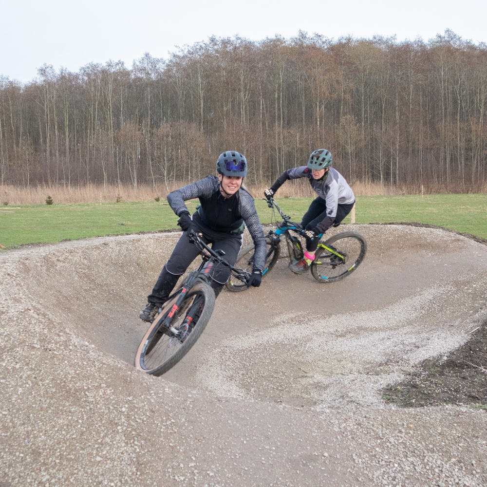 Twee mountainbikers met helm rijden achter elkaar door een kombocht op een gravelparcours in de buitenlucht, met bos op de achtergrond