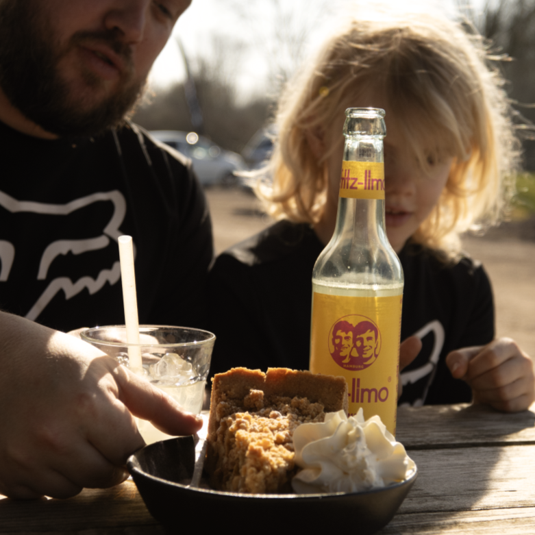 Vader en kind aan tafel op het zonnige terras van Indoor Mountainbike in Almere, met een stuk taart met slagroom en een koude limonade