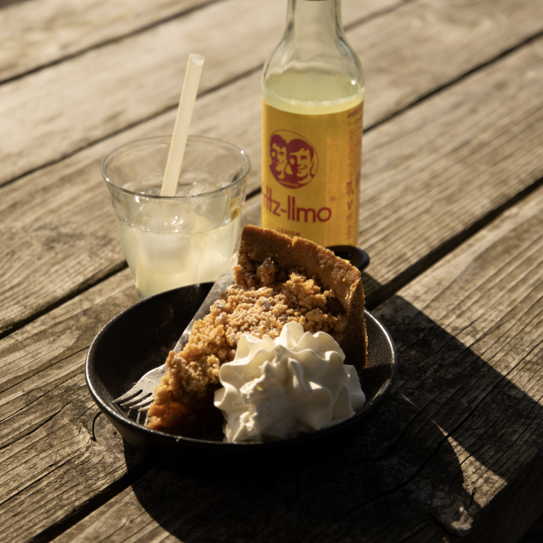 Stuk appeltaart met slagroom en een glas citroenlimonade op een houten picknicktafel in de zon