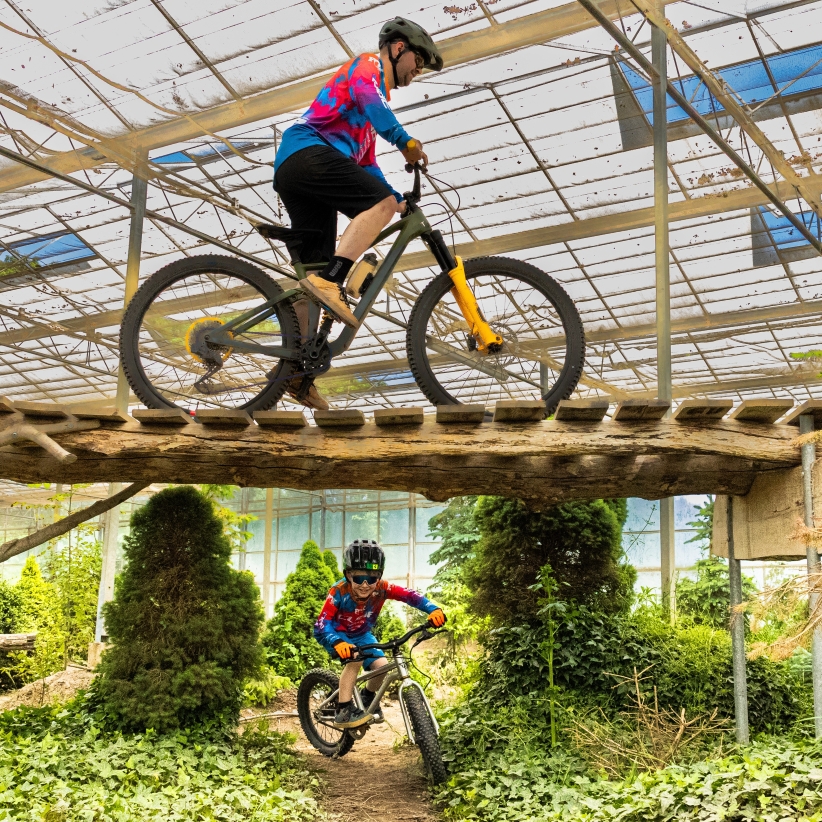 Volwassene rijdt op een mountainbike over een houten brug terwijl een kind eronderdoor fietst, tussen het groen in het overdekte parcours van Indoor Mountainbike Almere