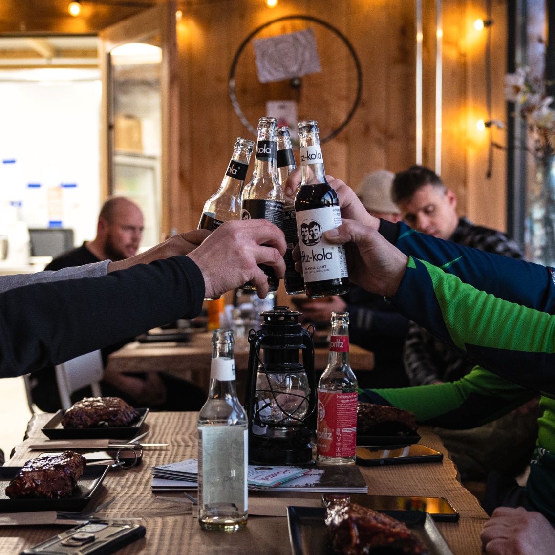 Groep proost met flesjes frisdrank boven een houten tafel met spareribs, in het sfeervol verlichte restaurant van Indoor Mountainbike Almere