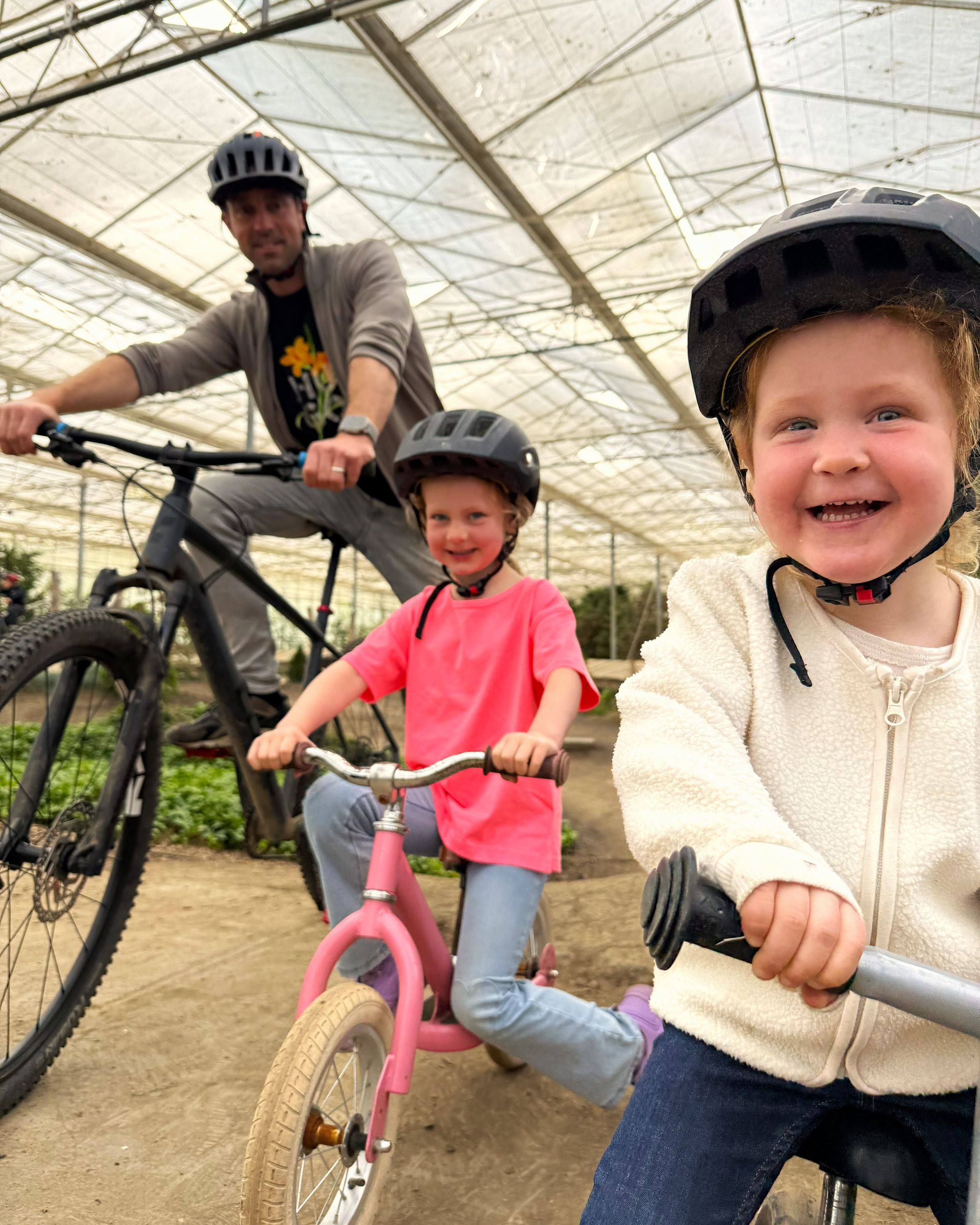 Twee lachende meisjes met helm op een loopfiets, met papa op de mountainbike erachter, op het indoor parcours van Indoor Mountainbike in Almere